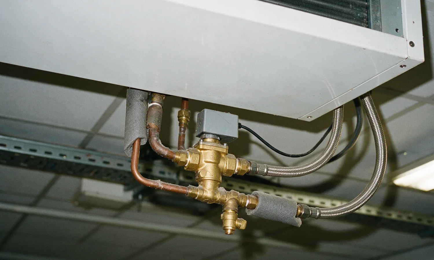 A brass valve body with a small grey electric actuator on top, hung under a white fan coil unit casing in an open ceiling void, with copper pipes, braided flexible hoses and insulation sleeves pushed back